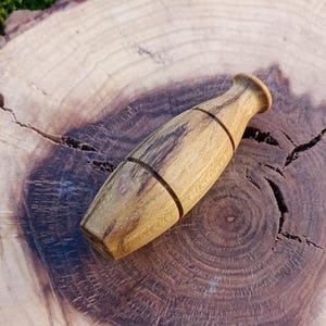 Könnte beinhalten: Ein Flaschenverschluss aus Holz mit einer natürlichen Holzmaserung. Der Verschluss ist in Form einer kleinen Vase geformt und hat ein dunkelbraunes Band um die Mitte.