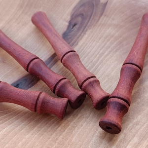 Könnte beinhalten: Vier handgefertigte Holzdübel mit rotbraunem Finish liegen auf einer hellen Holzoberfläche. Jeder Dübel hat eine zylindrische Form mit geschnitzten Ringen, die ein dekoratives Design bilden. Die Dübel sind wahrscheinlich für Bastelarbeiten oder Holzbearbeitung gedacht.