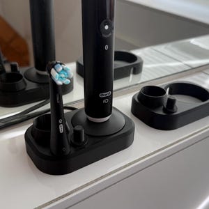 Könnte beinhalten: Eine schwarze elektrische Zahnbürste, die auf einer weißen Arbeitsplatte aufgeladen wird. Die Zahnbürste hat eine schwarz-weiße Ladestation und einen schwarzen Bürstenkopf. Die Zahnbürste ist mit "Oral-B iO" beschriftet.