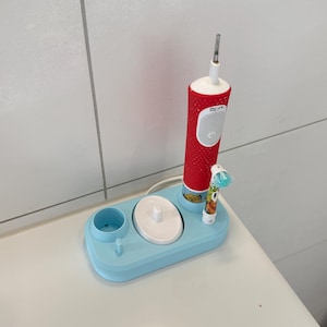 Könnte beinhalten: Eine rote elektrische Zahnbürste mit weißem Einschaltknopf und weißer Ladestation. Die Basis ist hellblau, oval geformt und verfügt über einen speziellen Steckplatz für einen kleineren Bürstenkopf mit buntem Aufdruck und blauen Borsten.