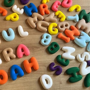 Könnte beinhalten: Eine Sammlung bunter, dreidimensionaler Buchstaben und Zahlen auf einer Holzoberfläche. Die Buchstaben bilden Namen wie "PAUL", "SARAH" und "NOAH". Die Buchstaben sind in verschiedenen Farben gehalten, darunter Rosa, Blau, Orange und Grün.