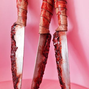 Puede incluir: Tres cuchillos de utilería con hojas plateadas y residuos texturizados de color rojo oscuro que simulan sangre a lo largo de los bordes. Cada mango está envuelto en material marrón claro con un patrón en espiral, formando un conjunto.