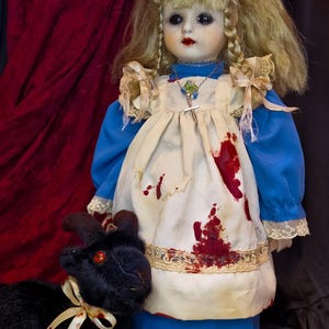 May include: A porcelain doll with blonde hair, wearing a blue dress and a blood-stained apron. A black stuffed animal with a red eye and a ribbon is next to the doll. The doll has a necklace with a cross pendant.