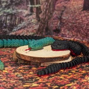 May include: Two toy snakes, one green and one black, are posed on a wooden log. The green snake has a red tongue sticking out. The black snake has a red stripe along its body.