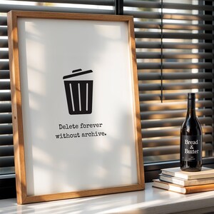 May include: A framed print with a wooden frame displays a black trash can icon and the text "Delete forever without archive." A bottle of "Bread & Butter" wine sits on a stack of books next to the frame.