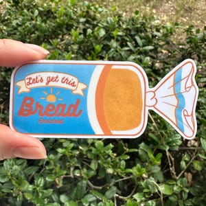 May include: A blue and orange sticker with the text "Let's get this Bread" and a sun graphic. The sticker is shaped like a loaf of bread in a wrapper.