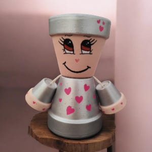 Könnte beinhalten: Handgefertigte Terrakotta-Blumentopfpuppe mit bemaltem Gesicht, silbernem Körper und rosa Herzakzenten. Die Puppe hat kleine Töpfe als Arme und sitzt auf einem Holzschemel. Der Hintergrund ist ein zartes Rosa.