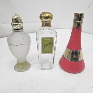 Pode incluir: Três frascos de perfume vintage. O frasco à esquerda é de vidro fosco com uma tampa dourada e o texto "COLOGNE SILK 3 FL OZ". O frasco do meio é de vidro transparente com uma tampa dourada e o texto "AVON Somewhere CREAM LOTION 4 FL OZ". O frasco à direita é uma garrafa rosa brilhante com uma tampa dourada e o texto "Ambush Perfume".