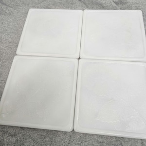 May include: Four square white ceramic coasters with a leaf design. The coasters have a slightly raised edge and a textured surface. The leaf pattern is visible on each coaster, adding a decorative element to the set.