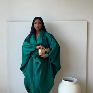 May include: A woman wearing a flowing, emerald green satin dress with a matching shawl. She holds a gold clutch. A large white vase is to the right. The background is a plain white wall.