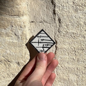 Op de afbeelding: Een ruitvormige geborduurde patch met een witte achtergrond, een zwarte rand en een reeks horizontale zwarte lijnen. Het ontwerp bevat verschillende zwarte zwaardsilhouetten die in verschillende richtingen over het oppervlak van de patch zijn geplaatst.