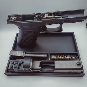 Puede incluir: Una pistola negra desmontada con varios componentes dispuestos en una bandeja negra. Los componentes incluyen el armazón, la corredera, el cañón, el muelle y la mira. La pistola se muestra sobre un fondo gris claro.