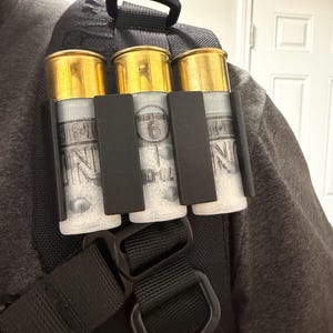 Op de afbeelding: Een zwarte shotgun shell houder bevestigd aan een rugzakriem. De houder bevat drie shotgun shells met goudkleurige toppen en doorzichtige hulzen gevuld met pellets. De shells worden op hun plaats gehouden door een zwarte plastic houder.