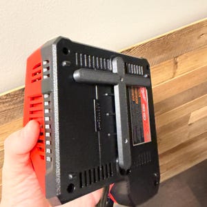 May include: A black and red power tool battery charger. The charger has a black body with red accents and ventilation slots. A black handle is attached to the back. A red and white label is visible on the back.
