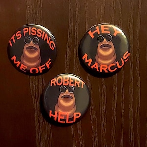 May include: Three black pinback buttons with a cartoon character wearing sunglasses. The buttons have orange text that reads "ITS PISSING ME OFF", "HEY MARCUS", and "ROBERT HELP". The cartoon character is brown with a large mouth.