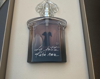 May include: A perfume bottle with a clear, ornate stopper and a dark, gradient-colored body. The bottle is in a box with a white interior. The text "La petite Robe noire" is written on the front of the bottle.