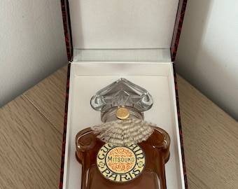 Può includere: Un flacone vintage di profumo Mitsouko di Guerlain in una scatola rossa e dorata. Il flacone è in vetro trasparente con un'etichetta dorata che dice "Mitsouko Paris". La scatola ha un design dorato con un cavallo e un cavaliere.