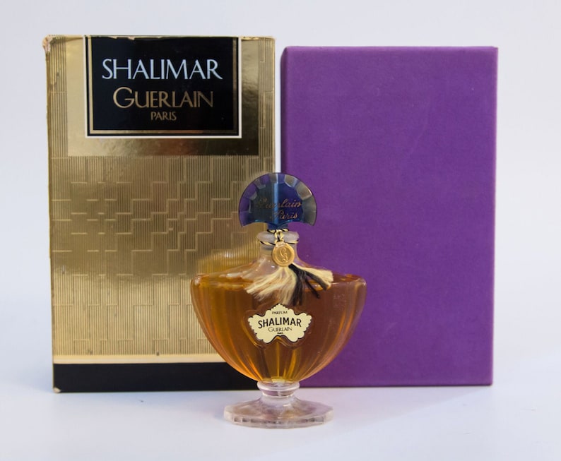 May include: A glass perfume bottle containing amber liquid with a fan-shaped blue stopper, a gold-toned tassel, and a label reading SHALIMAR GUERLAIN. It is displayed with a gold box featuring the text SHALIMAR GUERLAIN PARIS and a solid purple box.