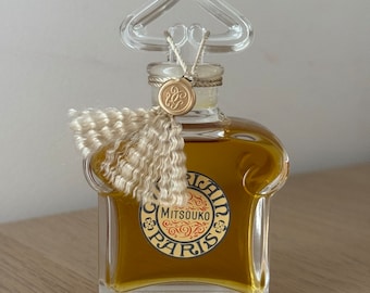 Può includere: Un flacone di profumo Mitsouko di Guerlain Paris in vetro trasparente. Il flacone ha una forma unica e curva e un tappo dorato con un fiocco. L'etichetta sul flacone presenta il nome del marchio e il nome del profumo in lettere dorate.