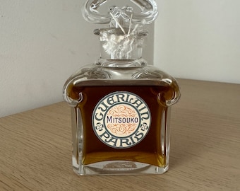 Può includere: Un flacone di profumo Mitsouko di Guerlain Paris in vetro trasparente. Il flacone ha un tappo a forma di cuore e un'etichetta con il nome del marchio e il nome del profumo.
