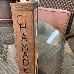 Könnte beinhalten: Ein rechteckiger Parfümflakon mit einem Holzdeckel und einer Holzfrontplatte. Auf der Platte steht vertikal das Wort "CHAMADE" und horizontal "HOMME". Die Flasche ist klar, und der Hintergrund ist unscharf.