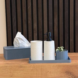 Può includere: Una mensola in legno con una scatola di fazzoletti, due contenitori cilindrici, un dosatore di sapone e una piccola pianta in un vassoio grigio. La mensola è appoggiata a una parete a doghe verticali marrone scuro e a una parete beige.