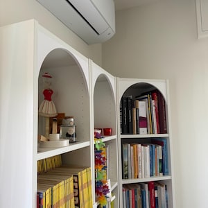 Op de afbeelding: Witte, gewelfde boekenplanken gevuld met boeken en decoratieve items. Een rood-witte jurkvorm, een pot en een lei zijn zichtbaar. De planken staan tegen een witte muur, met een airconditioner erboven.