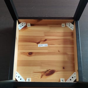 Peut inclure: Le dessous d'une chaise présentant un cadre de siège en bois clair naturel avec des grains et des nœuds visibles. Quatre pieds noirs sont fixés aux coins avec des équerres métalliques triangulaires blanches et des vis argentées.