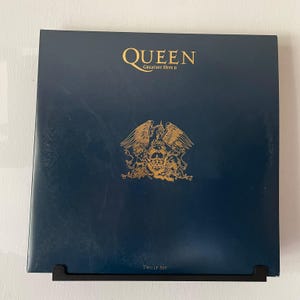 Könnte beinhalten: Ein quadratisches Vinyl-Schallplattencover für Queen Greatest Hits II. Das Cover ist dunkelblau mit goldener Schrift und einem goldenen Wappen mit Löwen und einem Phönix. Der Text lautet Queen Greatest Hits II oben und Two LP Set unten.