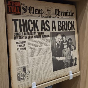 Può includere: Una copertina dell'album dei Jethro Tull intitolata "Thick as a Brick", progettata come un giornale, "The St. Cleve Chronicle & Linwell Advertiser", datato gennaio 1972. L'album è tenuto su uno scaffale da clip bianche.