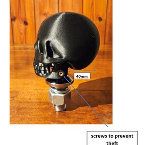 May include: A black skull-shaped object, approximately 40mm wide, mounted on a metal base with a bolt and nut. The design includes screws to prevent theft. The skull has detailed features.