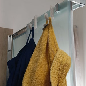 Può includere: Due asciugamani appesi a una porta doccia in vetro con ganci di plastica bianca. Un asciugamano è giallo e l'altro è blu.