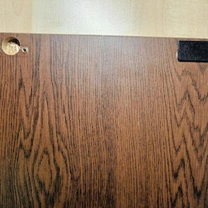 Può includere: Primo piano di una porta di armadio in legno marrone con un piccolo rettangolo nero sul lato destro e un foro rotondo sul lato sinistro.