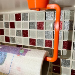 Può includere: Porta rotolo di carta da cucina in plastica arancione montato su un armadio bianco con un rivestimento in piastrelle a mosaico rosso e argento.