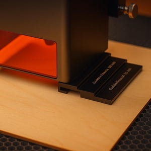 Puede incluir: Una grabadora láser negra con una ventana teñida de naranja, apoyada sobre una superficie de madera clara. La máquina tiene un botón de ajuste metálico y una base negra con texto grabado.