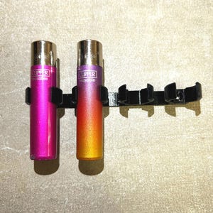 Può includere: Un porta accendini in metallo nero con due accendini. Entrambi gli accendini sono del marchio Clipper e sono riutilizzabili. Un accendino è rosa e l'altro è arancione.