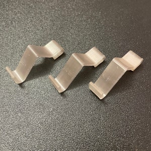 Op de afbeelding: Drie zilveren metalen clips, elk met een golvend ontwerp, zijn in een rij op een donkergrijs, getextureerd oppervlak gerangschikt.