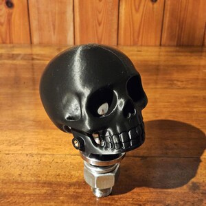 May include: A black skull-shaped gear shift knob. The skull has detailed features, including eye sockets, a nose cavity, and a full set of teeth. It is mounted on a metal base with a bolt and nut.