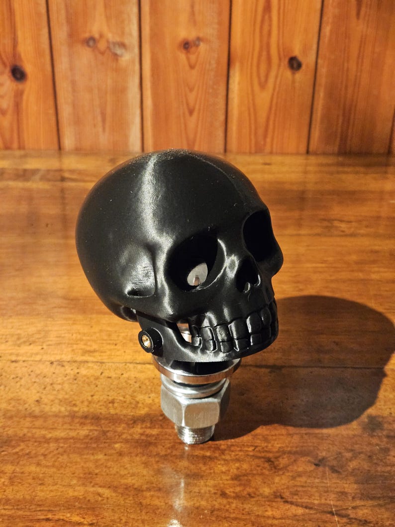 May include: A black skull-shaped gear shift knob. The skull has detailed features, including eye sockets, a nose cavity, and a full set of teeth. It is mounted on a metal base with a bolt and nut.