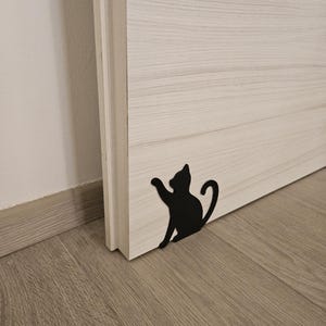 Op de afbeelding: Een zwarte metalen katvormige deurstopper staat aan de voet van een lichtgekleurde houten deur. De deurstopper is een silhouet van een kat met zijn poot omhoog. De deur en de vloer zijn van hout.