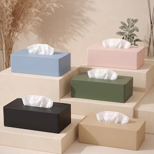 Könnte beinhalten: Eine Auswahl rechteckiger Taschentuchboxen in verschiedenen Farben: Hellblau, Rosa, Olivgrün, Schwarz und Beige. Jede Box enthält weiße Taschentücher, die aus der Oberseite herausragen. Die Boxen sind auf einer gestuften Präsentation angeordnet, modernes Wohnambiente.