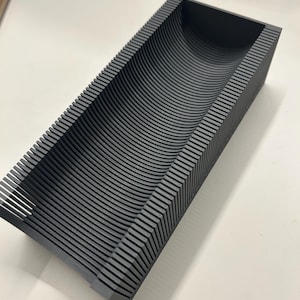Peut inclure: Un plateau rectangulaire gris foncé avec un intérieur incurvé. Le plateau est construit à partir de lignes parallèles superposées, créant une texture nervurée. La conception présente une auge profonde et arrondie, idéale pour organiser de petits objets.