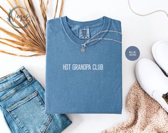 May include: A blue jean colored t-shirt with the text "HOT GRANDPA CLUB" in white. The shirt is folded and accessorized with a silver necklace, blue jeans, and white sneakers. A dried palm leaf and a beige scarf are also in the image.