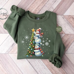 May include: A green sweatshirt with a Christmas tree and three snowmen on it. The snowmen are stacked on top of each other and are wearing hats and scarves. The tree is decorated with ornaments and a star on top. The sweatshirt is labeled "Military Green".