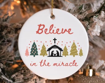 May include: White ceramic Christmas ornament with the words "Believe in the miracle" in red script. The ornament features a black nativity scene, a cross, and stylized Christmas trees in green and gold.