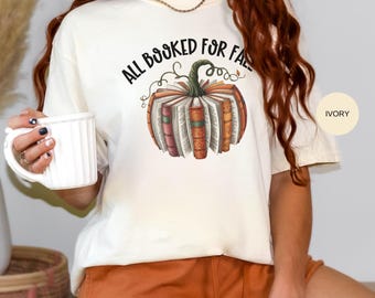 May include: Ivory-colored t-shirt with a pumpkin design made of books and the text "ALL BOOKED FOR FALL". The shirt is paired with orange shorts. A white mug is held in the hand.