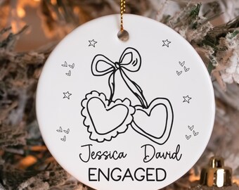 May include: A white ceramic ornament with a gold string for hanging. The ornament features a black line drawing of two hearts tied with a bow, surrounded by stars and hearts. The names "Jessica David" and the word "ENGAGED" are written in black script.