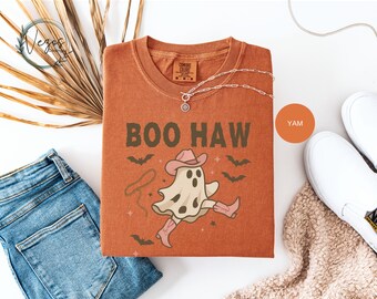 May include: A burnt orange t-shirt with the words "BOO HAW" in green, featuring a cartoon ghost wearing a cowboy hat and boots. The shirt is surrounded by bats and a lasso design. The shirt is paired with blue jeans and white sneakers.