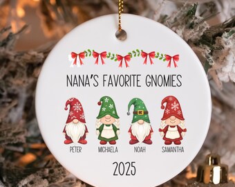 May include: A white ceramic Christmas ornament with the text "NANA'S FAVORITE GNOMIES" and the year 2025. Four gnome illustrations are below the text, each with a name. The ornament has a gold string for hanging.