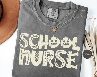 May include: A gray Comfort Colors t-shirt with the words "SCHOOL NURSE" in a light beige color. The design includes pumpkin and snake graphics. A silver necklace with a pendant is draped across the shirt.